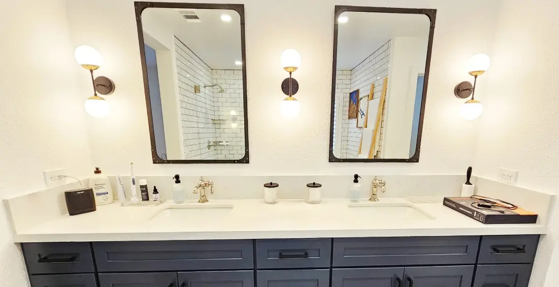 Double sink vanity with blue painted shaker style cabinets, white quartz top, polished nickel faucets, and black accessories. One framed mirror above each sink with sconces on either side.