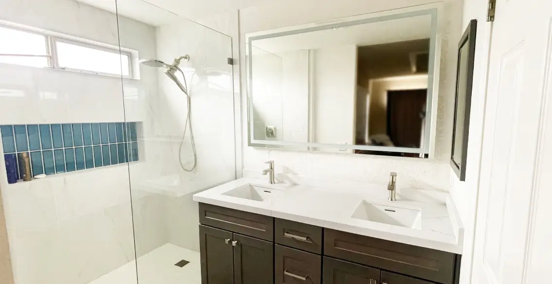 Tiled shower with large horizontal niche, blue decorative tile inside. White and grey tile on walls, white pebble on pan. Double sink vanity with dark maple stained cabinets, white quartz top, and single handle lav faucets. Brushed nickel fixtures.