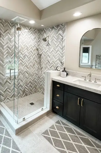 Large custom tiled shower with brown marble herringbone mosaic on wall, 2" x 2" mosaic on pan. Dark brown stained wood cabinets with taupe quartz top and polished nickel fixtures.