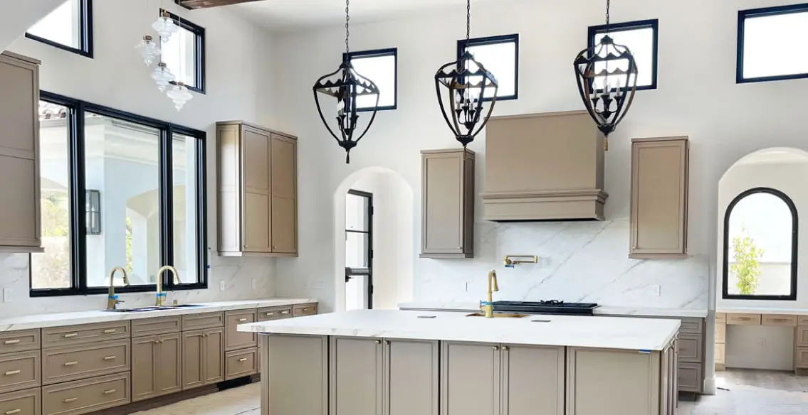 Large open concept kitchen with taupe painted modified shaker cabinets. Large custom wood hood over 48" range. Large island with Porcelain tops and 3 black pendant lights hanging above. Gold faucets and accents.