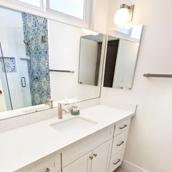 Vanity area with white shaker cabinets, quartz countertop, and brushed nickel fixtures. Shower shown in mirror reflection has tile on walls and frameless glass enclosure.