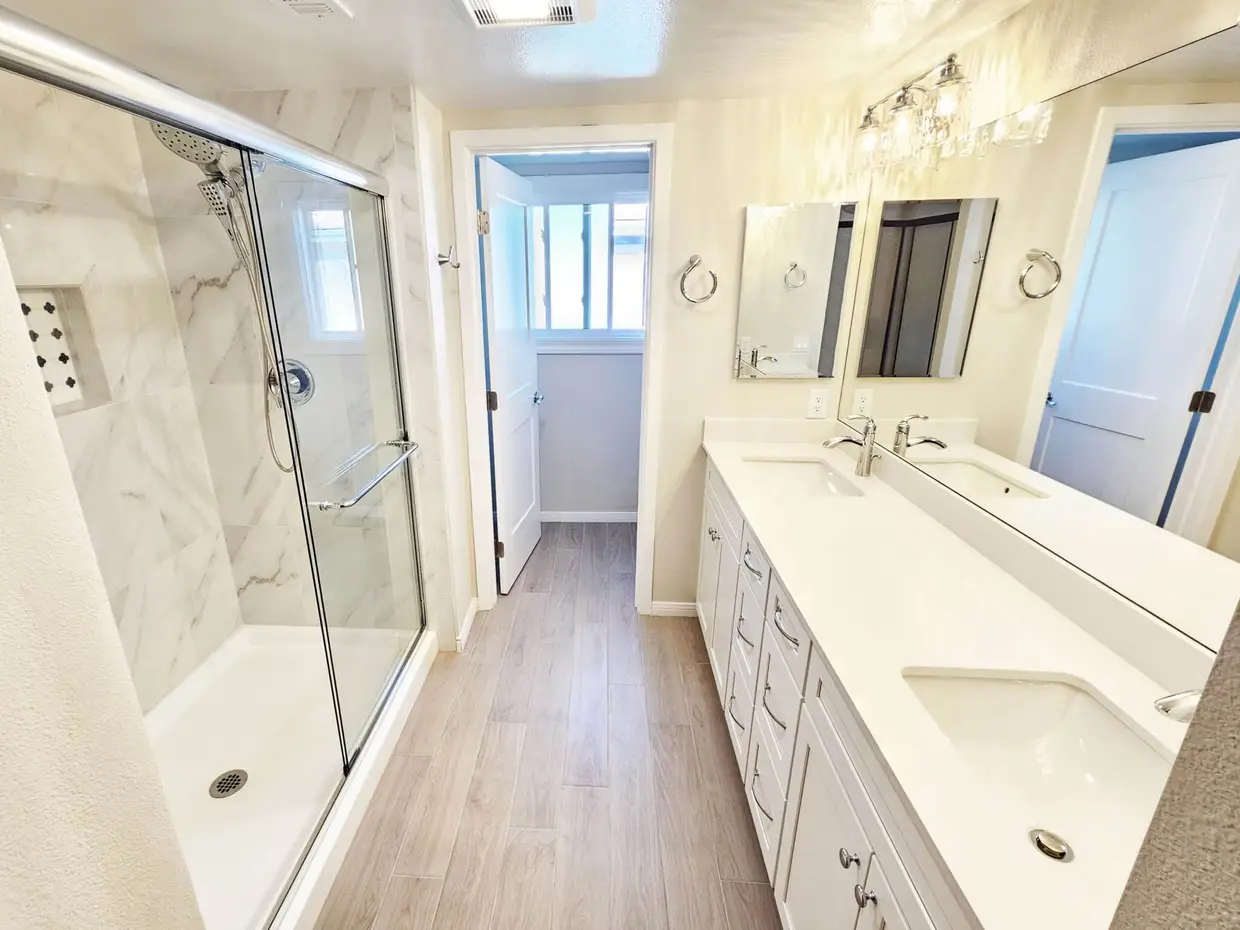 Modern bathroom remodel with double vanity, glass walk-in shower, marble style tile walls, and wood-look flooring.